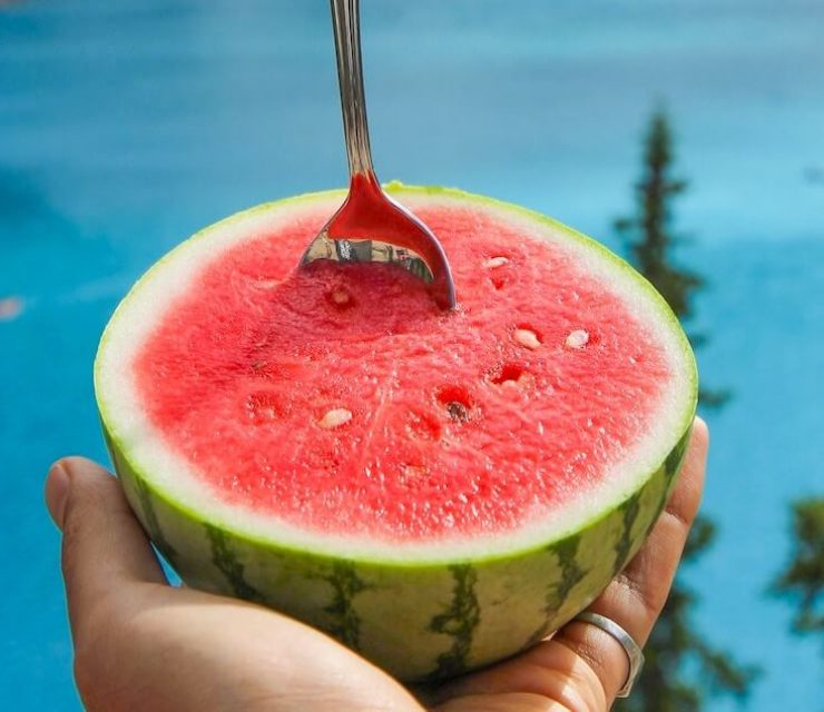 person holding watermelon with spoon in the fruit with backdrop of moraine lake
