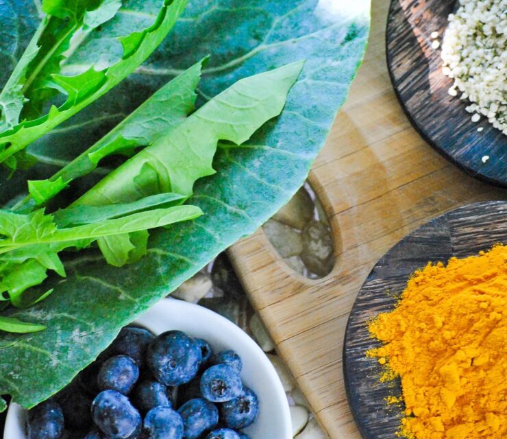 Health collage of leafy greens and super foods including collard greens, dandelion greens hemp seeds, antioxidant blueberries and anti-inflammatory turmeric on stones and a rustic wood cutting board.