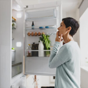 Side view of mature female with short hair standing in front of opened refrigerator at home with puzzled pensive facial expression, thinking of cooking breakfast, looking for ingredients