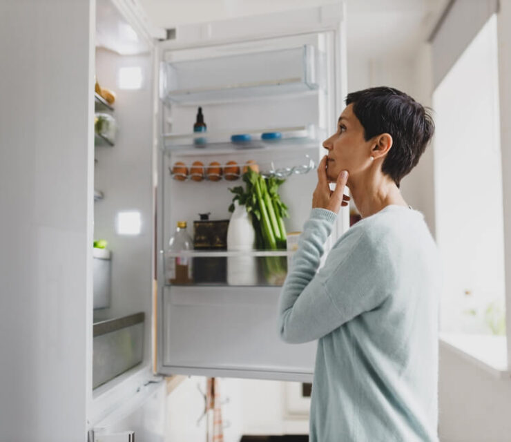 Side view of mature female with short hair standing in front of opened refrigerator at home with puzzled pensive facial expression, thinking of cooking breakfast, looking for ingredients