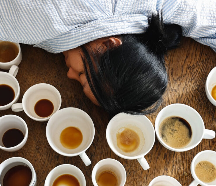 Exhausted, a tired female falls asleep at her working desk, surrounded by many empty coffee mugs, indicating a problem with caffeine addiction in female worker.