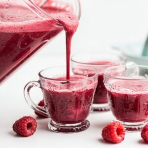 Vibrant raspberry smoothie being poured from a blender into small vintage glass cups, thick rich deep red color with visible texture and light foam, fresh raspberries scattered nearby