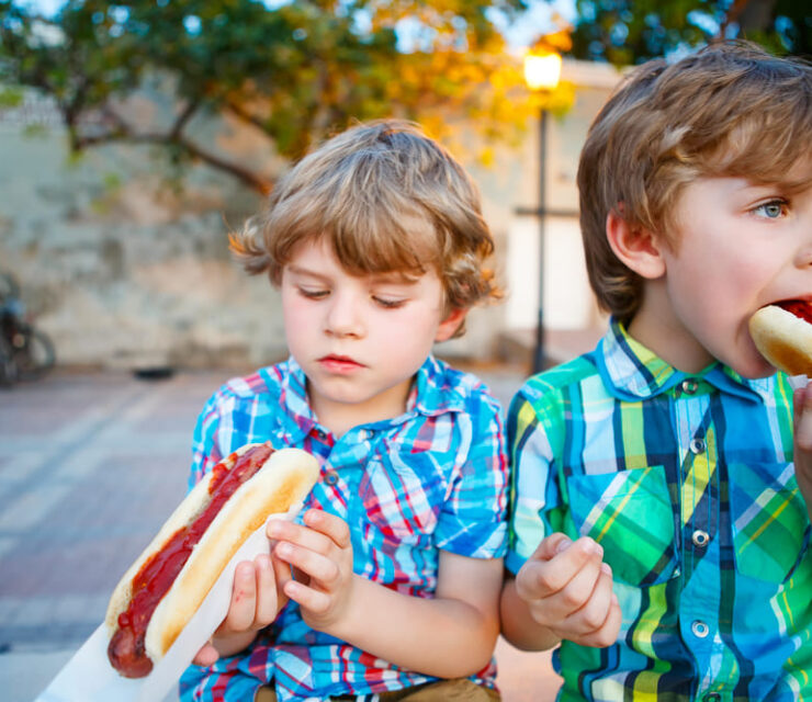 Two little kid boys eating hot dogs outdoors. Siblings enjoying their meal. Hotdog as nhealthy food for children.