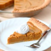 slice of pumpkin pie on a white plate with full pie in the background on wooden table
