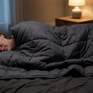 Ultra-realistic nighttime lifestyle photograph of a woman sleeping under a heavy weighted blanket, clearly showing the distinctive characteristics of a true weighted blanket. The blanket is thick, dense, and visibly compressed against her body, conforming tightly to her torso and arms due to gravity. Subtle stitched square compartments (weighted blanket quilting) are visible, with gentle downward pull and realistic pressure distribution. The blanket appears heavier than a normal duvet, with minimal loft and a structured, weighted drape.